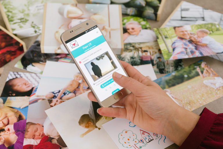 Main qui tient un téléphone. L'application La personne télécharge une photo sur l'application Mamizette. Photos imprimées et disposées en arrière plan.